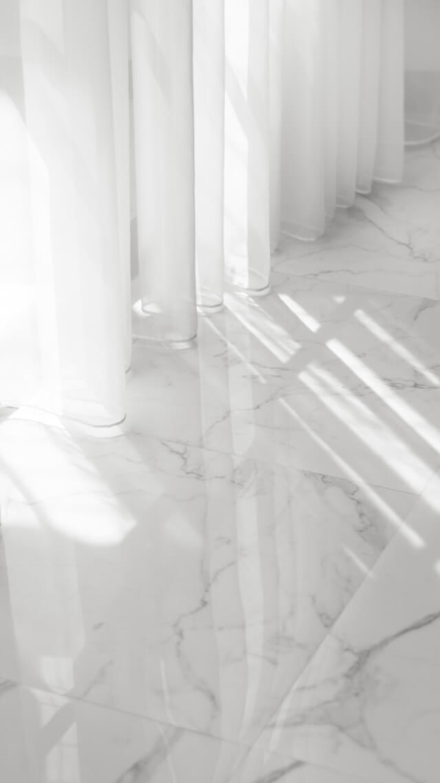 Black and white image of light shining through curtains onto a marbled floor.